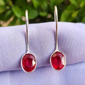 Puede incluir: Un par de pendientes colgantes con piedras preciosas facetadas de color rojo intenso en forma ovalada, engastadas en metal plateado pulido. Cada pendiente tiene un gancho de alambre plateado largo y fino que se extiende hacia arriba.