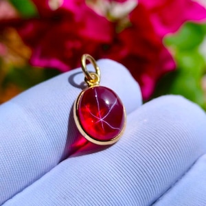 Könnte beinhalten: Ein runder, tiefroter Cabochon-Anhänger in einer polierten, goldfarbenen Metallfassung mit einer kleinen runden Öse an der Oberseite. Der Edelstein zeigt ein ausgeprägtes sechsstrahliges Sternmuster auf seiner glatten, durchscheinenden Oberfläche.