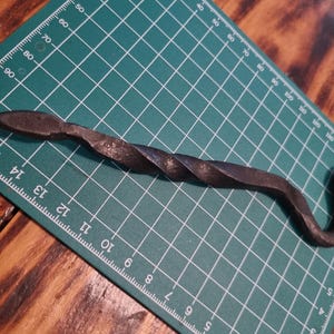 May include: A dark grey, hand-forged metal snake figurine with a twisted body texture. The object is positioned on a green cutting mat with white grid lines and numerical markings, showing a length of approximately 16.5 cm.