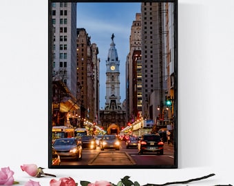 May include: Framed print of a city street scene at dusk. The image features a tall, ornate building with a clock tower at the end of a street lined with buildings. Cars and buses are visible on the road.
