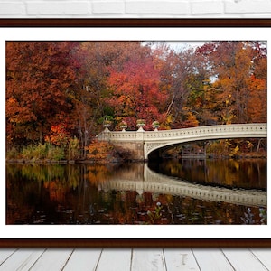 May include: A framed photograph of a scenic autumn landscape. A white arched bridge spans a dark lake, reflecting the colourful foliage of the trees. The trees display vibrant hues of red, orange, and yellow, creating a picturesque scene.