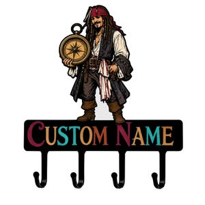 Puede incluir: Un perchero de pared con un personaje pirata sosteniendo una brújula sobre un panel horizontal negro con el texto CUSTOM NAME en letras multicolores y cuatro ganchos de metal negro en el borde inferior.
