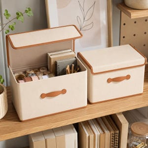 May include: Two rectangular beige storage boxes with brown trim and matching brown handles. The larger box is open, revealing notebooks, pens, and various spools of ribbon inside. The smaller box is closed, sitting beside the open one.