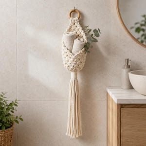 May include: A cream-coloured macrame wall hanging basket featuring a circular wooden ring at the top and a long, thick tassel at the bottom. The woven pouch contains two folded light-coloured towels and a small sprig of green eucalyptus leaves.