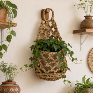 Op de afbeelding: Een beige, geweven jute plantenhanger met een geknoopt, open netontwerp, opgehangen aan een houten wandhaak. De hanger bevat een witte pot met een groene klimop die met ranken uit de getextureerde, vezelige structuur naar buiten groeit.