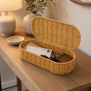Puede incluir: Una cesta de almacenamiento ovalada de color amarillo mostaza con una tapa abatible unida. La cesta contiene un sobre blanco, un par de gafas de sol con montura oscura y un pequeño objeto de cuero marrón para organizar objetos.