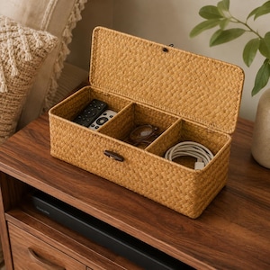 May include: A rectangular woven storage box with a hinged lid and a dark brown wooden toggle closure. The interior is divided into three sections, containing two black remote controls, a pair of eyeglasses, and a coiled white charging cable.