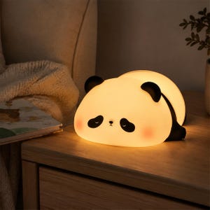 May include: A glowing panda-shaped night light emitting a warm, soft yellow illumination. The lamp features a smooth, rounded body with black ears, black patches around the closed eyes, a small nose, and subtle pink blush marks on the cheeks.