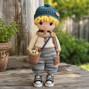 May include: A handmade crochet doll with yellow hair wearing a blue beanie, a cream-coloured long-sleeve shirt with brown buttons, and blue and white striped trousers. The doll carries a small brown crossbody bag containing a tiny stuffed animal.