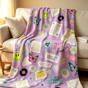 May include: A light purple fleece blanket featuring a repeating pattern of stickers including yellow smiley faces, colorful butterflies, daisies, hearts, vinyl records, lightning bolts, stars, and handwritten notes with the text Violet, Youth, and Pain Dreamer.
