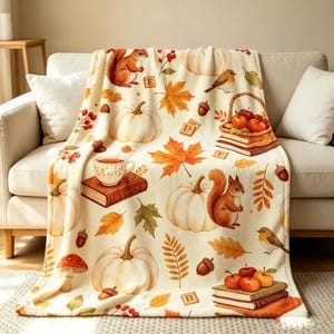 May include: A cream-colored throw blanket featuring an autumn-themed pattern of orange squirrels, white pumpkins, red apples in baskets, brown acorns, colorful maple leaves, mushrooms, birds, books, and small wooden letter tiles with the letters E, P, F, and B.