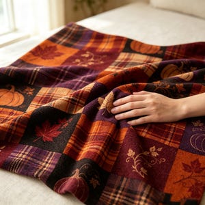 K&ouml;nnte beinhalten: Eine weiche Wohndecke mit einem Patchwork-Design aus herbstlichen Quadraten. Das Muster zeigt orangefarbene K&uuml;rbisse, rote Ahornbl&auml;tter und braune Karomuster in verschiedenen Schattierungen von Dunkellila, gebranntem Orange und Dunkelbraun.