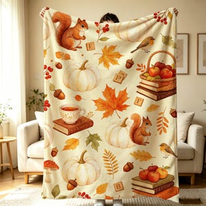 May include: A cream-colored throw blanket featuring an autumn-themed pattern of orange squirrels, white pumpkins, red berries, brown acorns, maple leaves, books, a teacup, a bird, and wooden blocks with the letters E, F, and B printed on them.