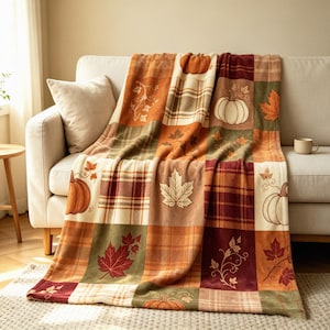 May include: A soft throw blanket featuring a patchwork design with alternating squares of autumn-themed patterns, including orange pumpkins, red and gold maple leaves, and brown plaid prints in a warm color palette of cream, orange, and deep red.