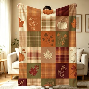 May include: A rectangular throw blanket featuring a patchwork design of autumn-themed squares. The pattern includes alternating blocks of plaid in shades of brown, orange, and green, interspersed with illustrations of pumpkins and maple leaves.