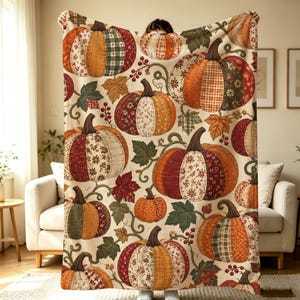 Op de afbeelding: Een crèmekleurige plaid met een herhalend patroon van pompoenen gemaakt van diverse patchworkstoffen in tinten oranje, rood, groen en bruin. Het ontwerp bevat geïllustreerde groene ranken, donkergroene bladeren en kleine rode bessen.