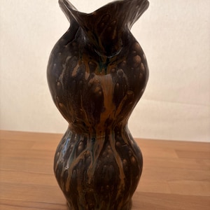 Könnte beinhalten: Eine Keramikvase mit ausgeprägter Sanduhrform und einem gewellten, ausgestellten Rand. Die Oberfläche hat eine dunkelbraune Grundfarbe mit vertikal verlaufenden Glasurmustern in Beige-, Kupfer- und dezenten Blautönen, die einen Effekt erzeugen.