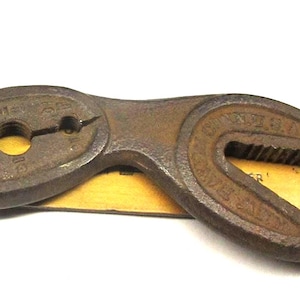 May include: A vintage, rusty brown multi-tool wrench with two curved ends and a central oval section with three holes. The wrench is lying on a yellow ruler, with the measurements in inches.