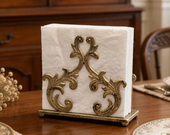 Puede incluir: Un servilletero de metal dorado con un diseño ornamental simétrico de volutas en la parte frontal. El soporte contiene una pila de servilletas de papel cuadradas de color blanco y se asienta sobre una base rectangular con pequeñas patas.