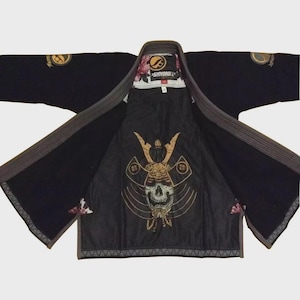 May include: A black martial arts gi jacket featuring a large graphic of a skull wearing a samurai helmet on the inner back panel. The interior lining displays a floral pattern, and the brand name Shoyoroll is printed on the neck label.
