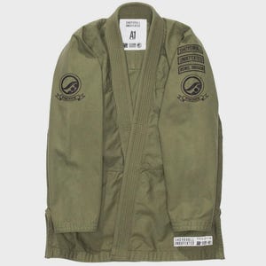May include: An olive green martial arts gi jacket featuring black circular logos on both sleeves. The left sleeve displays stacked text reading Shoyoroll, Undefeated, and Rebel Division. A white label at the neck shows the size A1 and brand text.