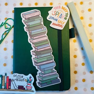 May include: A dark green notebook featuring a tall sticker of stacked books, a sticker with the text THE WAY YOU SPEAK TO YOURSELF MATTERS, and a sticker with the text Stay and read. A green paperclip and a light gray pen are placed nearby.