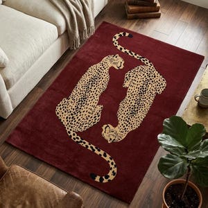 Op de afbeelding: Een rechthoekig vloerkleed in een diepe bordeauxrode kleur met twee beige cheeta's met zwarte vlekken in een gespiegelde, symmetrische opstelling. De cheeta's zijn zittend afgebeeld met hun lange staarten die naar buiten krullen.