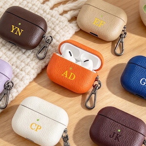 Puede incluir: Una colección de siete estuches texturizados para auriculares en varios colores, incluyendo marrón oscuro, beige, naranja, lavanda, azul, blanco y granate. Cada estuche presenta un monograma de dos letras, un pequeño punto verde y un clip de metal negro.