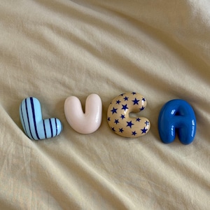 May include: Four individual decorative letters spelling LUCA. The L is light blue with dark blue vertical stripes. The U is solid pale pink. The C is beige with a pattern of small dark blue stars. The A is a solid, vibrant royal blue.