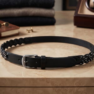 Peut inclure: Une ceinture en cuir noir avec un motif tressé sur les côtés, ornée de petits clous métalliques argentés. La ceinture présente une section en cuir lisse à l'avant avec une boucle en métal argenté et un passant en cuir assorti.