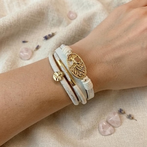 May include: A multi-strand white leather bracelet featuring a gold-toned oval charm with a tree design and a small gold-toned circular charm. The bracelet is constructed with several layers of white leather cord and secured with gold-toned metal hardware.