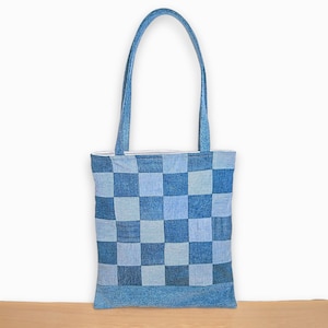 May include: A rectangular tote bag constructed from denim fabric featuring a patchwork design of alternating light and dark blue squares. The bag includes two long shoulder straps made of the same blue denim material and a solid blue base panel.