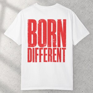 May include: A white short-sleeved t-shirt featuring the text BORN DIFFERENT printed in a large, distressed red font across the center. The shirt has a crew neckline and a standard fit, showcasing a clean design with bold, weathered lettering.