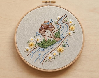 May include: A circular wooden embroidery hoop containing a beige fabric panel with a hand-stitched design. The embroidery depicts a small mushroom character wearing a hat, paddling a green leaf boat down a blue stream, surrounded by white daisies.