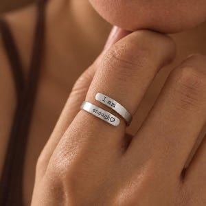 Può includere: Un anello aperto color argento con due estremità. L'estremità superiore è incisa con il testo I am e l'estremità inferiore con il testo enough seguito da un piccolo simbolo a forma di cuore, creando un aspetto sovrapposto.