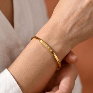May include: A gold-toned metal bangle bracelet featuring a smooth, polished finish. The exterior surface of the bracelet is engraved with the letters K and M separated by a small heart symbol. The bangle has a slim, uniform band design.