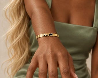 May include: A gold-toned metal cuff bracelet featuring a cutout design of three bears in a row, with one large bear followed by two smaller bears, centered on the smooth, polished surface of the band.