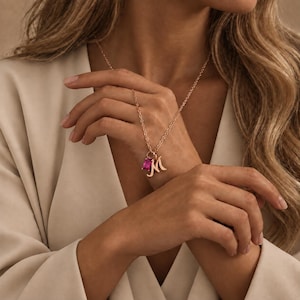 May include: A rose gold chain necklace featuring two charms: a rectangular, faceted hot pink gemstone and a cursive letter M pendant. The necklace is worn against a light beige garment, showcasing the metallic finish and the vibrant pink stone.