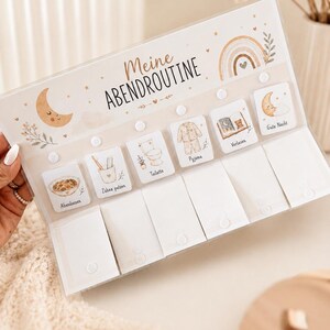 Op de afbeelding: Een rechthoekig avondroutinebord van acryl met de titel Meine Abendroutine en een afbeelding van een maan en regenboog. Het bevat zes witte vierkante kaarten met illustraties en Duitse tekst, elk bevestigd met een witte drukknoopsluiting.
