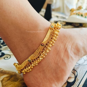 May include: A gold-toned anklet featuring a wide, textured chain band adorned with small, dangling spherical charms. The design includes tiny, colorful floral accents in red, green, and black enamel along the length of the metallic chain.