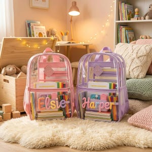 May include: Two clear backpacks, one pink and one lavender, each featuring a front pocket with a personalised name patch reading Elsie and Harper respectively. The backpacks are filled with colourful notebooks, pencils, and various school supplies.