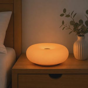 Puede incluir: Una lámpara de mesa circular en forma de rosquilla con un acabado blanco mate suave que emite un brillo naranja cálido, suave y difuso. La lámpara presenta un centro hueco y se asienta sobre una superficie plana para iluminación.