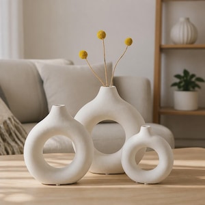 May include: A set of three white ceramic vases with a circular, donut-shaped design and a narrow neck. The central vase contains three thin stems topped with small, round, yellow flowers. The vases have a smooth, matte finish and flat bases.