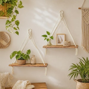 Peut inclure: Deux étagères rectangulaires en bois suspendues par des cordes en macramé blanc cassé avec des boucles circulaires et des pompons. Les étagères supportent une petite plante verte, un vase en céramique beige, un cadre et un petit panier tressé.