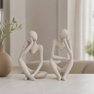 Könnte beinhalten: Zwei abstrakte, menschenähnliche Skulpturen aus einem hellbeigen Material mit gesprenkelter, steinartiger Textur. Jede Figur ist in einer sitzenden, nachdenklichen Pose dargestellt, wobei eine Hand am Gesicht und ein Arm auf dem Knie ruht.