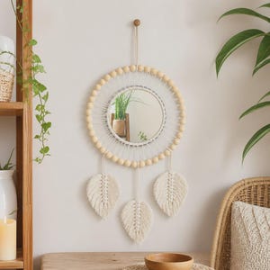 May include: A circular wall mirror framed by light-colored wooden beads and white macrame cord. The mirror features three hanging macrame leaf-shaped tassels at the bottom and is suspended by a single cord from a small wooden wall hook.