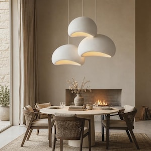 Puede incluir: Un conjunto de tres lámparas colgantes de color blanco mate con formas de cúpula orgánicas y curvas suspendidas del techo. Cada luminaria presenta un exterior liso y una parte inferior que emite una luz cálida sobre una mesa de comedor redonda.