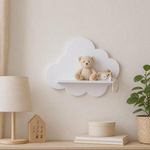 May include: A white wall-mounted shelf shaped like a cloud. The shelf holds a light brown plush teddy bear sitting upright and a small wooden toy camera with a strap. The cloud-shaped shelf is mounted on a plain wall, providing a flat surface for display.