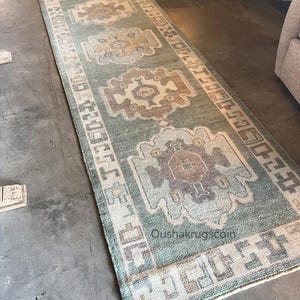 May include: A long rectangular runner rug featuring a sage green background with four repeating geometric medallions in shades of beige, brown, and cream. The border displays a repeating block pattern in cream and brown. Text reads Oushakrugscoin.
