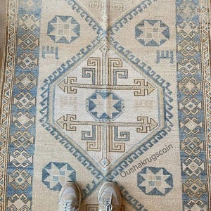 May include: A rectangular area rug featuring a beige background with a central diamond motif in blue and tan. The design includes geometric patterns, star-like shapes, and a border with intricate repeating motifs. The text Oushakrugscoin is visible.